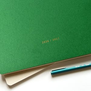 May include: A green notebook with the gold text "2026 / 2027" printed on the cover. A teal pen rests on the corner of the notebook. The notebook is open, revealing a cream-colored page.
