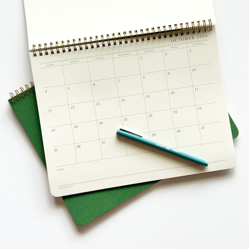 May include: An open spiral-bound calendar for October 2026, with a teal pen resting on the page. The calendar is on top of a green notebook. The calendar grid is visible, with days of the week and dates clearly marked.