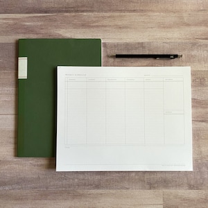 K&ouml;nnte beinhalten: Ein w&ouml;chentlicher Terminplaner mit einem gr&uuml;nen Notizbuch auf einer Holzoberfl&auml;che. Der Planer hat einen Bereich f&uuml;r jeden Tag der Woche mit Platz f&uuml;r Notizen und Termine. Der Planer ist auf wei&szlig;em Papier mit schwarzen Linien gedruckt.