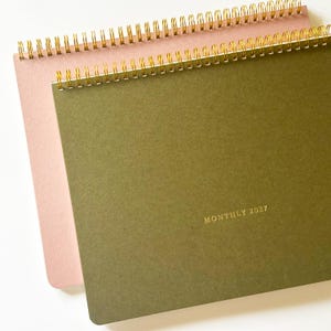 May include: Two spiral-bound landscape planners with gold wire binding. The top planner has a solid olive green cover with the gold-stamped text "MONTHLY 2027" in the bottom right corner. The bottom planner has a solid light pink cover.