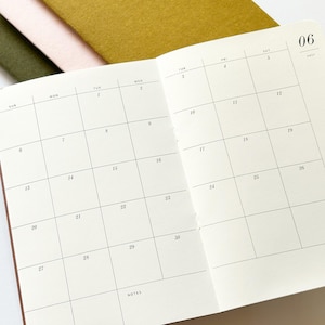 May include: An open planner showing a monthly calendar grid for June, labeled 06, with numbered days from 1 to 30 and a section for notes. The planner has off-white pages with black text and is stacked atop three other notebooks with felt covers.