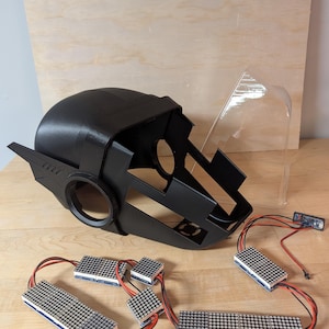 May include: Black, angular helmet with a clear visor, likely for cosplay or a costume. Several small LED matrix displays are connected with wires, suggesting an electronic component. The items are arranged on a light-colored wooden surface.