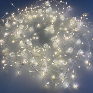 May include: A string of warm white LED lights with small, clear, crackled glass globe accents. The lights are arranged in a tangled, circular pattern, creating a soft, ambient glow. Suitable for decorative lighting.