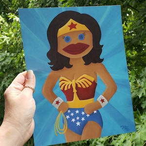 May include: A whimsical painting of a Wonder Woman sock puppet. The artwork features a blue background with radiating light beams. The puppet has a red and gold bodice, blue shorts with white stars, and a gold lasso. The eyes are blue buttons.