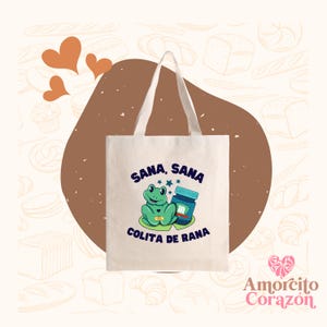 May include: A cream-colored canvas tote bag featuring a graphic of a green frog with a bandage on its chest, sitting next to a blue jar. The text "SANA, SANA COLITA DE RANA" is printed in dark blue letters above and below the frog illustration.