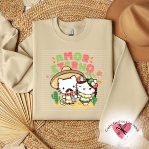 May include: Beige sweatshirt featuring a cartoon couple, one with a mustache and sombrero, the other with a flower crown. The text "AMOR ETERNO" is above them. The sweatshirt is on a woven surface with a brown hat.