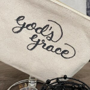 May include: A beige canvas zippered pouch with the words "God's grace" in black script. A silver watch and black beaded bracelet are visible in the foreground.