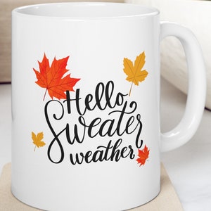 Puede incluir: Taza de cerámica blanca con un gráfico en blanco y negro que dice "Hello Sweater Weather". El gráfico está rodeado de hojas de arce naranja y amarillo.