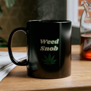 Op de afbeelding: Zwarte keramische mok met een wit ontwerp met de tekst "Weed Snob" en een groen marihuanablad. De mok is gevuld met een warme drank en er stijgt stoom op uit de bovenkant.