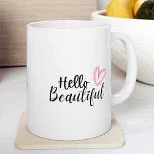 May include: White ceramic mug with a black cursive inscription that reads "Hello Beautiful" and a pink heart design. The mug is sitting on a beige coaster.
