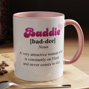 May include: White ceramic mug with a pink rim and handle. The mug features a pink graphic with the word "Baddie" in a retro font and the definition "[bad-dee] Noun A very attractive woman who is constantly on Fleek and never ceases to slay."
