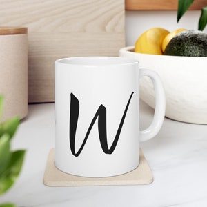 May include: White ceramic mug featuring a large black letter "W". The mug is placed on a beige coaster. A wooden board and a bowl of fruit are visible in the background. The mug is a standard size.