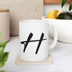 Puede incluir: Taza de cerámica blanca con una letra "H" negra escrita a mano en la parte delantera. La taza está sobre un posavasos beige.