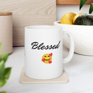 May include: White ceramic coffee mug with a black cursive inscription "Blessed" and a yellow smiley face with red hearts. The mug is sitting on a beige coaster.