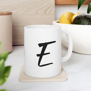 May include: White ceramic mug with a black handwritten letter "E" on the front. The mug is sitting on a beige coaster.