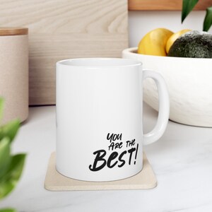 May include: White ceramic coffee mug with a black inscription that reads "You are the Best!" The mug is sitting on a beige coaster.