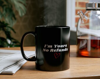 May include: Black coffee mug with white text that reads "I'm Yours No Refunds" with a red heart. The mug is on a wooden table with a newspaper and a glass teapot in the background.