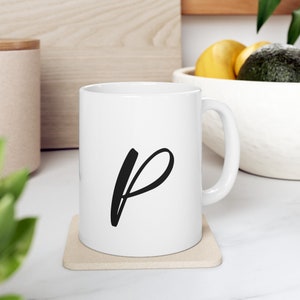 Può includere: Tazza in ceramica bianca con una lettera "P" in corsivo nero sul davanti. La tazza è appoggiata su un sottobicchiere beige.