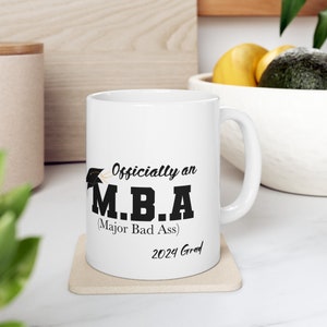 Puede incluir: Taza de cerámica blanca con un gráfico negro que dice "Officially an MBA (Major Bad Ass)" y "2024 Grad".