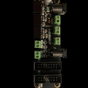 May include: A black circuit board featuring electronic components such as capacitors, resistors, and connectors. The board includes green terminal blocks, black connectors, and the text "A 15-20VDC". The background is black.