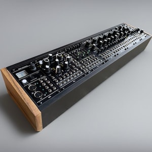 May include: A black synthesizer with numerous knobs, buttons, and ports. The wooden side panels contrast with the black faceplate. The device is designed for electronic music production and performance, featuring a variety of controls for sound manipulation.