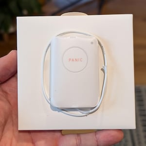 Puede incluir: Un dispositivo electrónico blanco con la palabra "PANIC" en un círculo rojo, sobre un cuadrado blanco. Un cable blanco está enrollado alrededor del dispositivo. El dispositivo está en la mano de una persona.