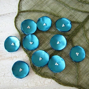 May include: A collection of ten teal blue fabric flower embellishments with white centers. The flowers are arranged on a brown burlap background.