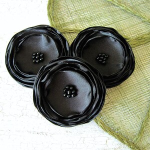 Puede incluir: Tres flores de tela de satén negro con centros de cuentas negras. Las flores están dispuestas sobre un fondo verde tejido.