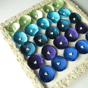 May include: A white frame with a gradient of green, blue, and purple fabric flowers. Each flower has a white pearl center.