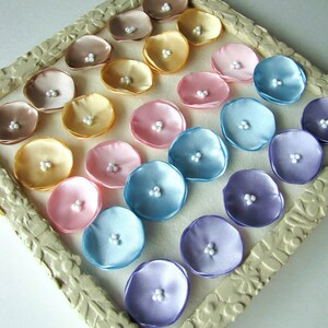 May include: A collection of 20 satin flower embellishments in various pastel colors, arranged in a circular pattern on a white background. The flowers are made of satin fabric and have a pearl-like center.
