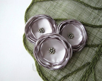 May include: Three large, layered, pale lavender satin flowers with silver bead centers on a green woven background.