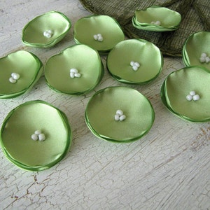 May include: A collection of 10 green satin flower petals with white pearl centers. The petals are arranged on a white textured surface.