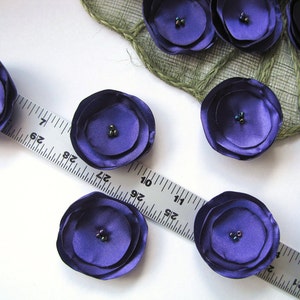 May include: Purple satin fabric flowers with a layered design and a small black bead in the center of each flower. The flowers are arranged on a white surface with a ruler.
