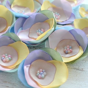 Puede incluir: Una colección de flores de tela de color pastel con cuentas de color perla en el centro. Las flores están superpuestas en tonos de amarillo, rosa, azul y morado.