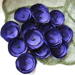 May include: A cluster of purple satin fabric flowers with iridescent bead centers. The flowers are arranged on a green burlap background.