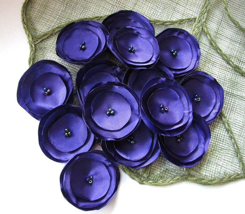 May include: A cluster of purple satin fabric flowers with iridescent bead centers. The flowers are arranged on a green burlap background.