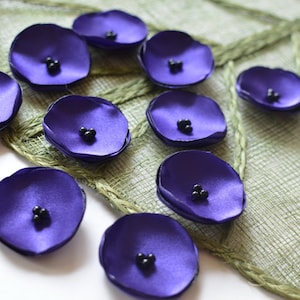 May include: Ten purple satin fabric flowers with black bead centers. The flowers are arranged on a green woven surface.
