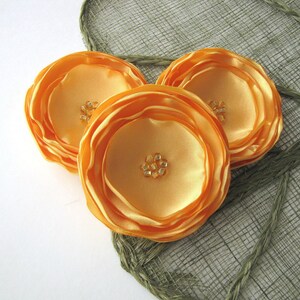 May include: Three orange fabric flower embellishments with a layered design. Each flower has a center made of small, clear rhinestones.