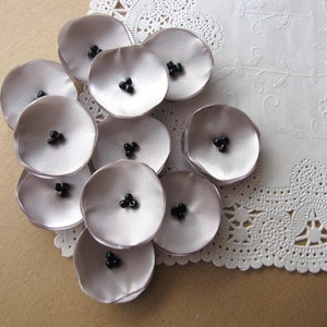 May include: A cluster of ten gray fabric flowers with black bead centers. The flowers are arranged in a circular pattern on a white doily.