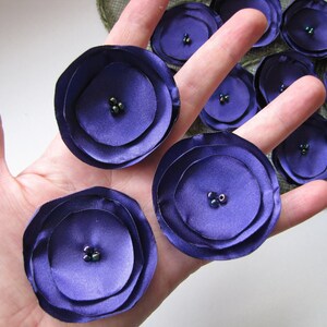 May include: Purple fabric flowers with multiple layers and dark green beads in the center. The flowers are on a hand.