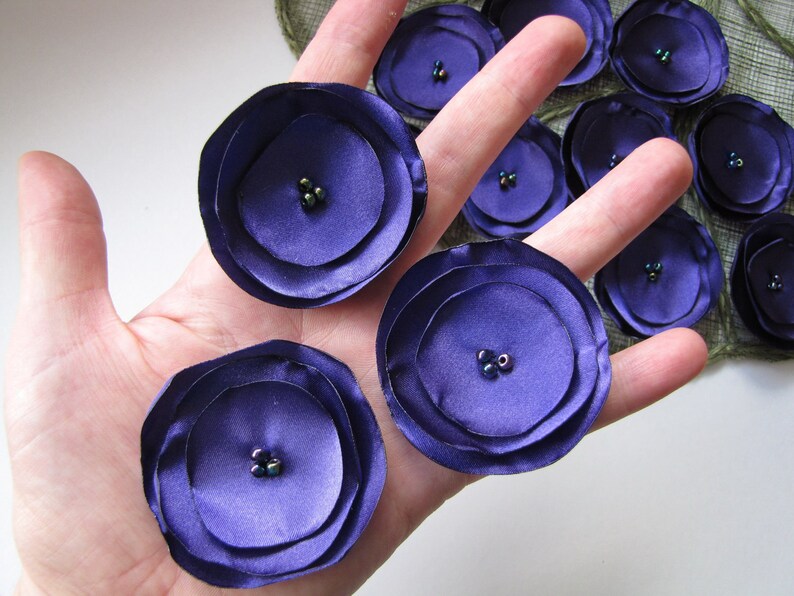 May include: Purple fabric flowers with multiple layers and dark green beads in the center. The flowers are on a hand.