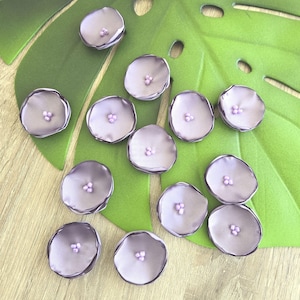 May include: A collection of lavender-colored fabric flower embellishments with small, light purple bead centers. The flowers are arranged on a large, green leaf-shaped cutout and a light wood-grain surface.