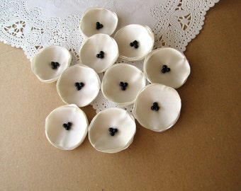 May include: A collection of white fabric flowers with black bead centers. The flowers are arranged on a brown surface.