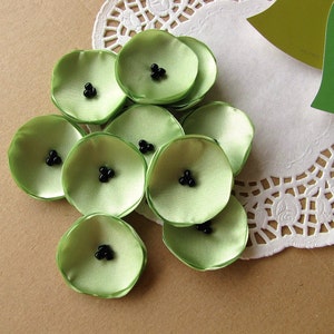 May include: A collection of green satin flower shapes with black beads in the center. The flowers are arranged on a white doily.