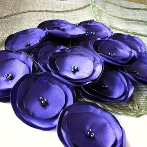 May include: A cluster of purple fabric flowers with iridescent beads in the center. The flowers are arranged in a circular pattern and are made of multiple layers of fabric.