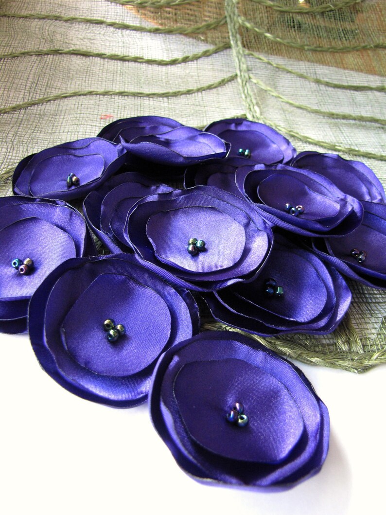 May include: A cluster of purple fabric flowers with iridescent beads in the center. The flowers are arranged in a circular pattern and are made of multiple layers of fabric.