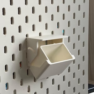 May include: White plastic wall-mounted storage bin with a small shelf on top. The bin is attached to a white pegboard.
