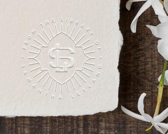 May include: A white embossed stamp with the text "From the Library of Samantha Leyland" and a monogram of the letters "S" and "L" in a circle with a sunburst design.
