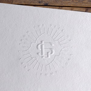 Op de afbeelding: Wit briefpapier met een reliëflogo. Het logo toont de initialen "SL" in een decoratief embleem, omringd door de tekst "FROM THE DESK OF SARA LYNNE". Het papier ligt op een houten achtergrond.