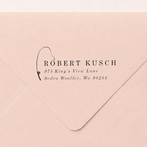 May include: A light pink envelope featuring a black ink return address stamp. The text reads ROBERT KUSCH, 975 King's View Lane, Sedro Woolley, Wa 98284, accompanied by a thin, black line drawing of a fishing rod curved over the name.